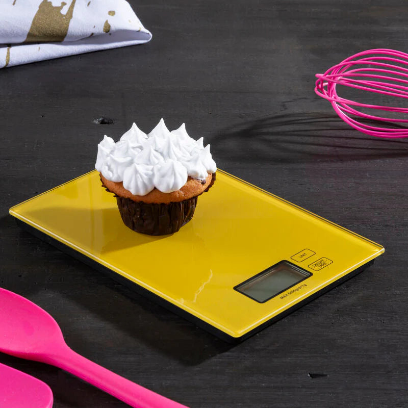 yellow digital scale with a cupcake on top of it sat on a black countertop surrounded by glipses of kitchen utensils.