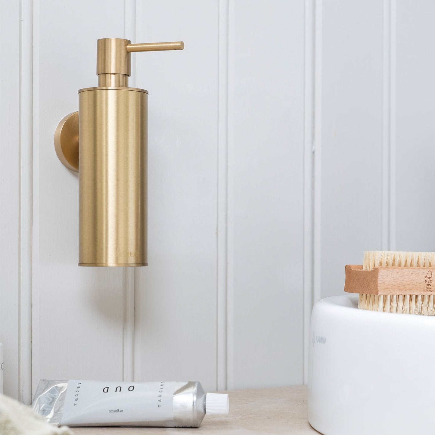 Gold soap dispenser on a white wall with a nail brush nearby and a white tube below.