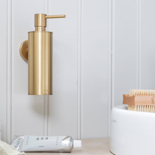Gold soap dispenser on a white wall with a nail brush nearby and a white tube below.