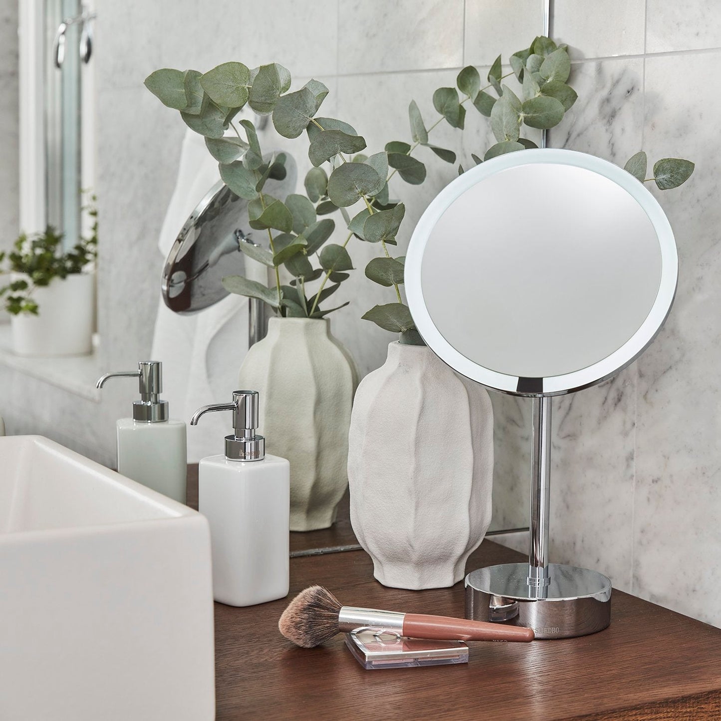 Makeup mirror and soap dispenser on a bathroom counter with toiletries and decor