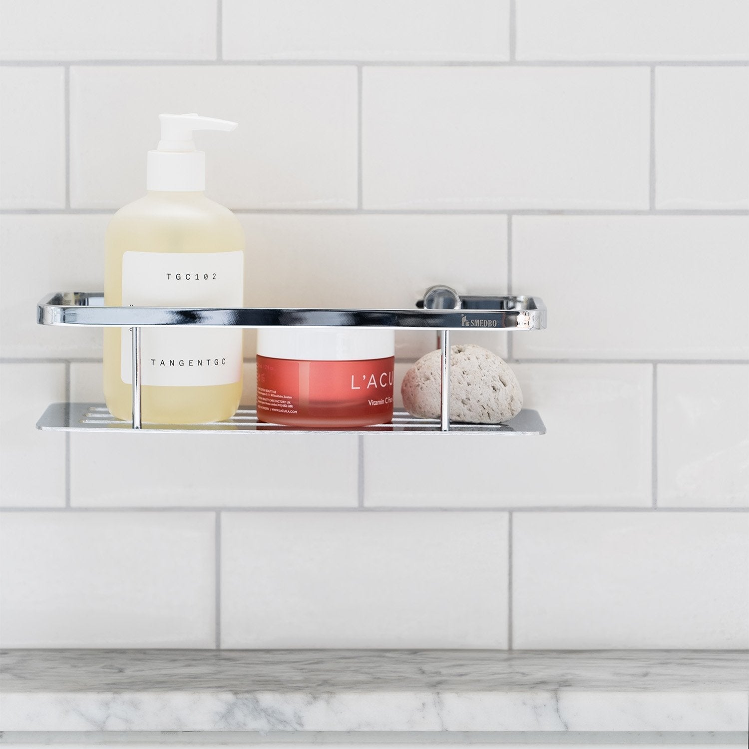 Smedbo Sideline DK3001 Polished Chrome Shower Basket — Close-Up of Solid Perforated Tray Styled with Pump Bottle, Skincare Jar and Pumice Stone on White Subway Tiles