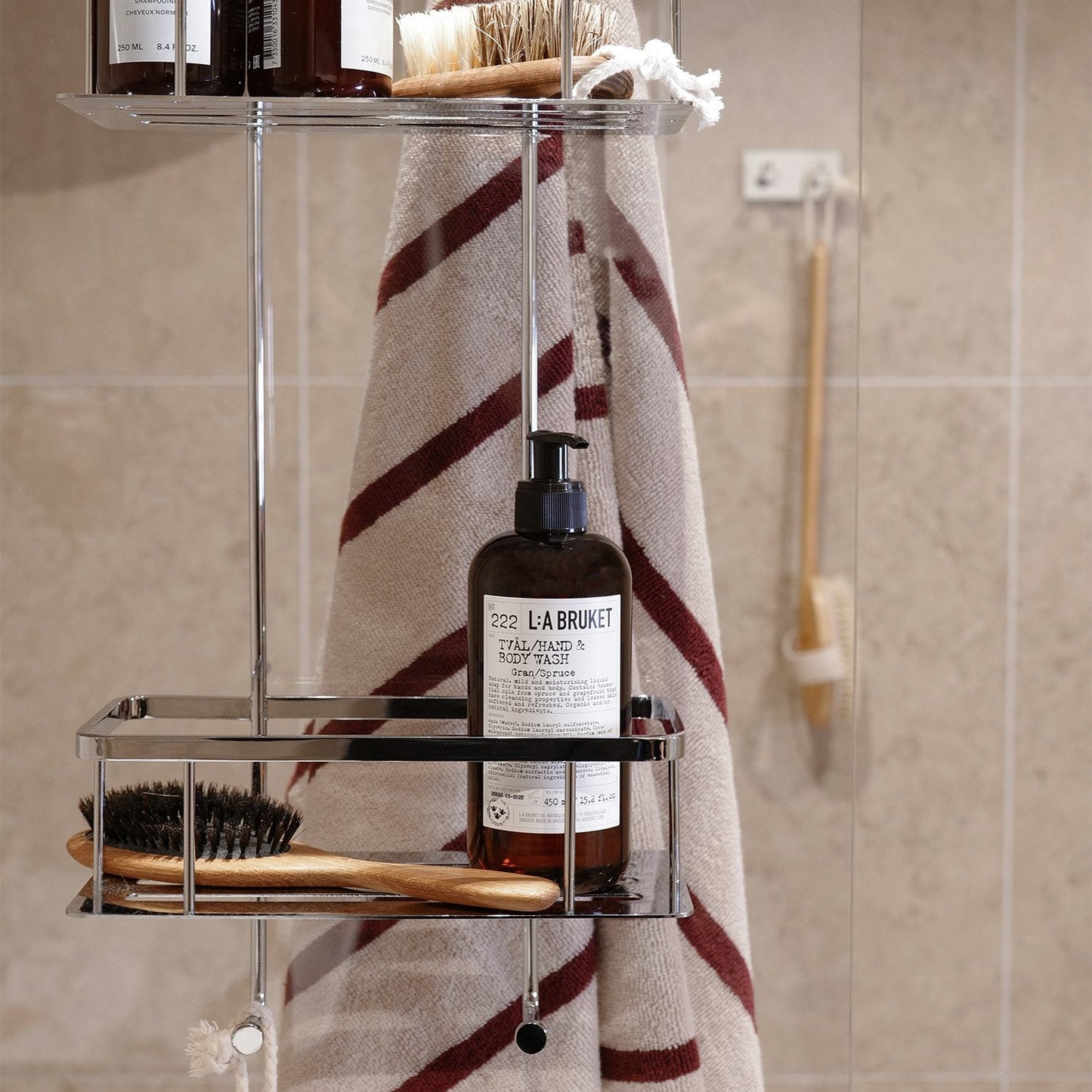 Smedbo Sideline DK3041 Hanging Shower Basket in Use — Striped Towel on Outward S-Hooks, L:A Bruket Body Wash and Wooden Brushes in Lower Tray, Beige Tiled Bathroom