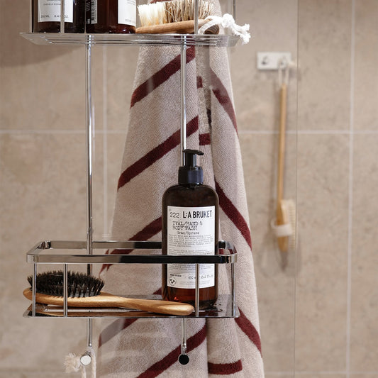 Smedbo Sideline DK3041 Hanging Shower Basket in Use — Striped Towel on Outward S-Hooks, L:A Bruket Body Wash and Wooden Brushes in Lower Tray, Beige Tiled Bathroom