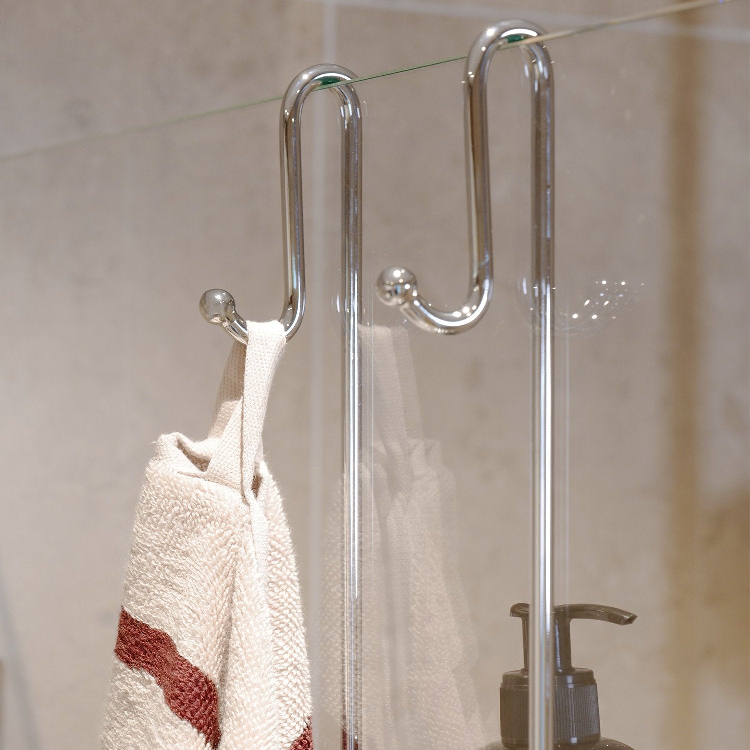 Smedbo Sideline DK3041 S-Hook Detail — Outward-Facing Towel Hook on Bathroom Side of Frameless Glass Panel, Striped Cream and Burgundy Towel Hanging, Pump Bottle Visible Inside Shower