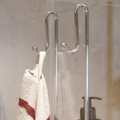 Smedbo Sideline DK3041 S-Hook Detail — Outward-Facing Towel Hook on Bathroom Side of Frameless Glass Panel, Striped Cream and Burgundy Towel Hanging, Pump Bottle Visible Inside Shower