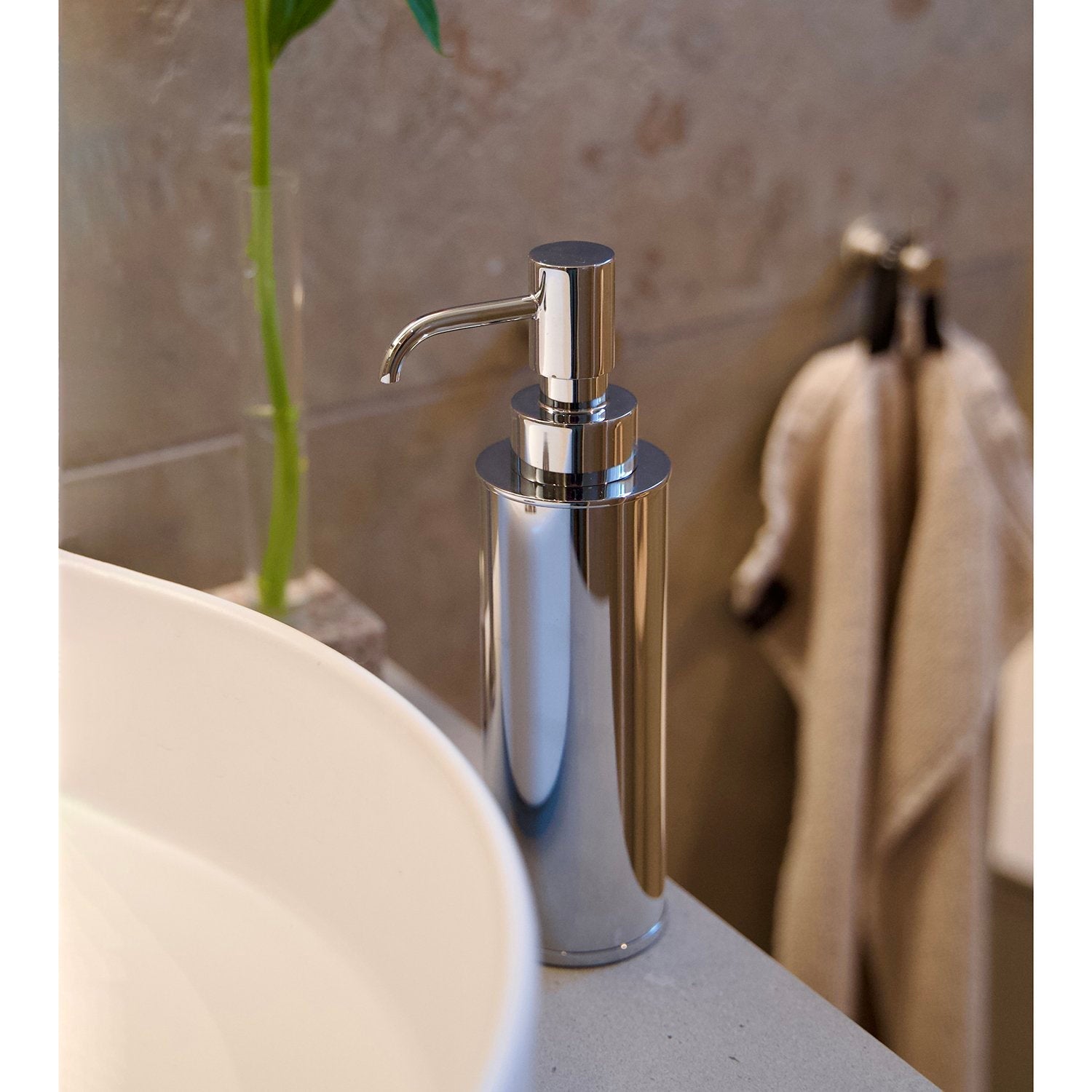 Smedbo Outline FK254 Polished Chrome Freestanding Soap Dispenser on White Stone Vanity Beside Round Countertop Basin — Bamboo Stem and Linen Towel in Background