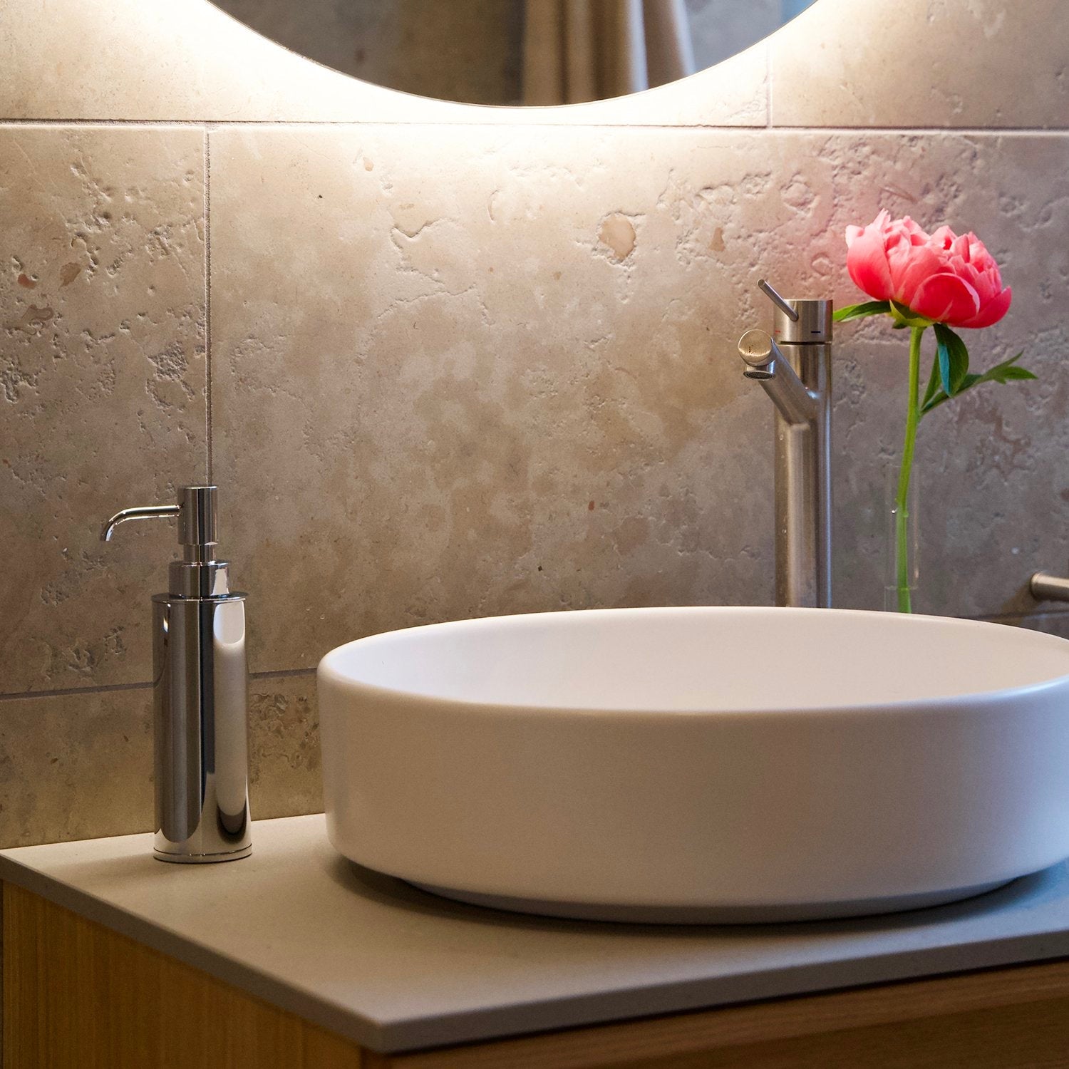 Smedbo Outline FK254 Polished Chrome Soap Dispenser on Oak Vanity — Round Countertop Basin, Chrome Mixer Tap, Pink Peony in Glass Vase, Travertine Tiles and Backlit Round Mirror